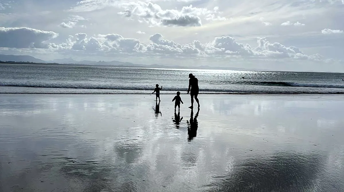 An adult and two small children at the water's edge at low tide, silhouetted against a bright cloudy sky, their reflections in the wet sand